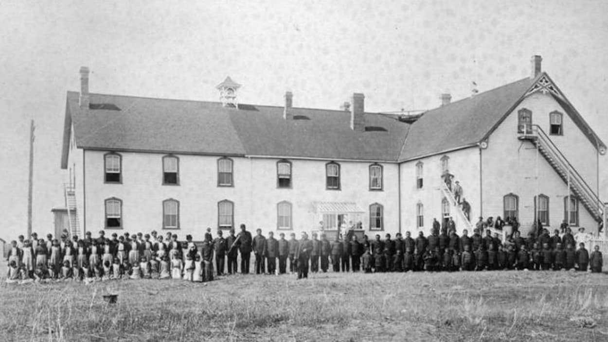 Des élèves debout devant le pensionnat autochtone Battleford Industrial School en 1895. Un édifice à deux étages.