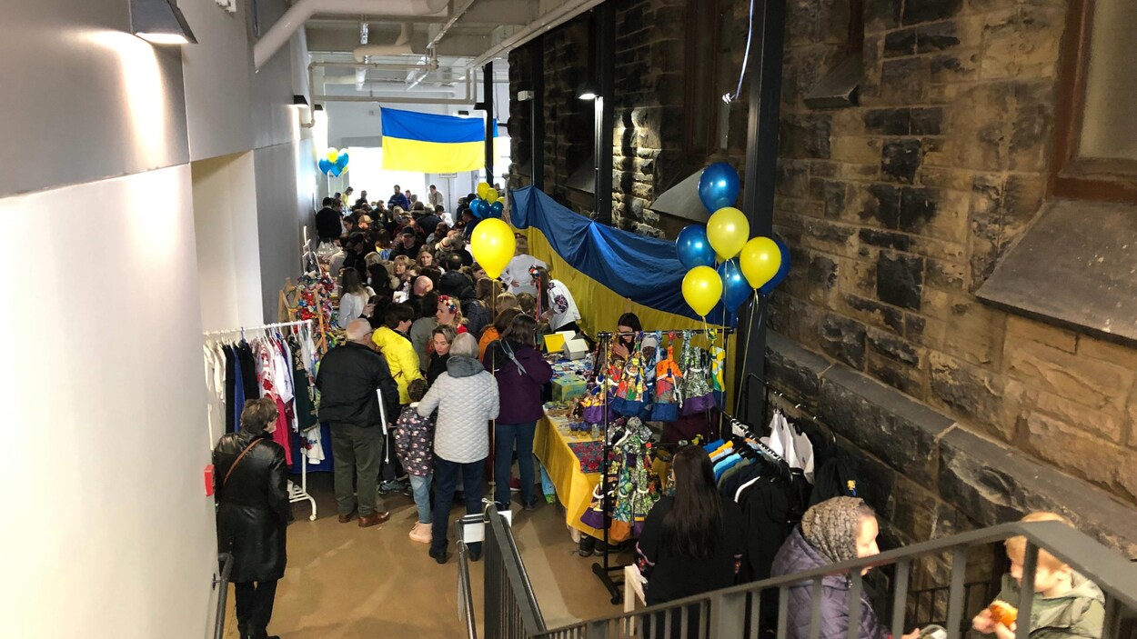 Vue d'une mezzanine, une foule se presse dans un couloir étroit rempli de kiosques de marchandises, décoré de drapeaux de l'Ukraine et de ballons bleus et jaunes gonflés à l'hélium.