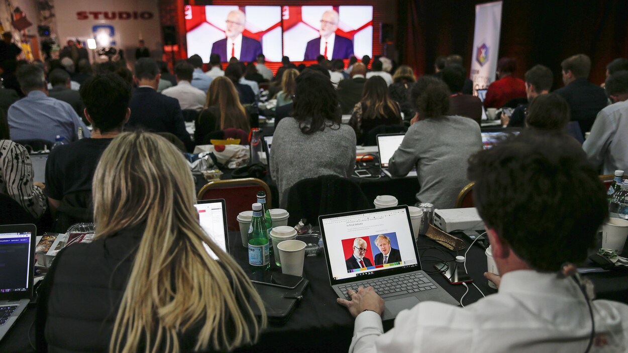 Des journalistes sont attablés, en rangées, avec leur ordinateur portable, devant des écrans géants sur lesqueon voit Jeremy Corbyn.