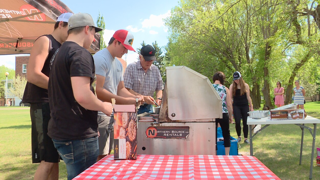 Quatre hommes s'activent autour du barbecue avec en arrière-plan quatre femmes qui discutent dans le parc.