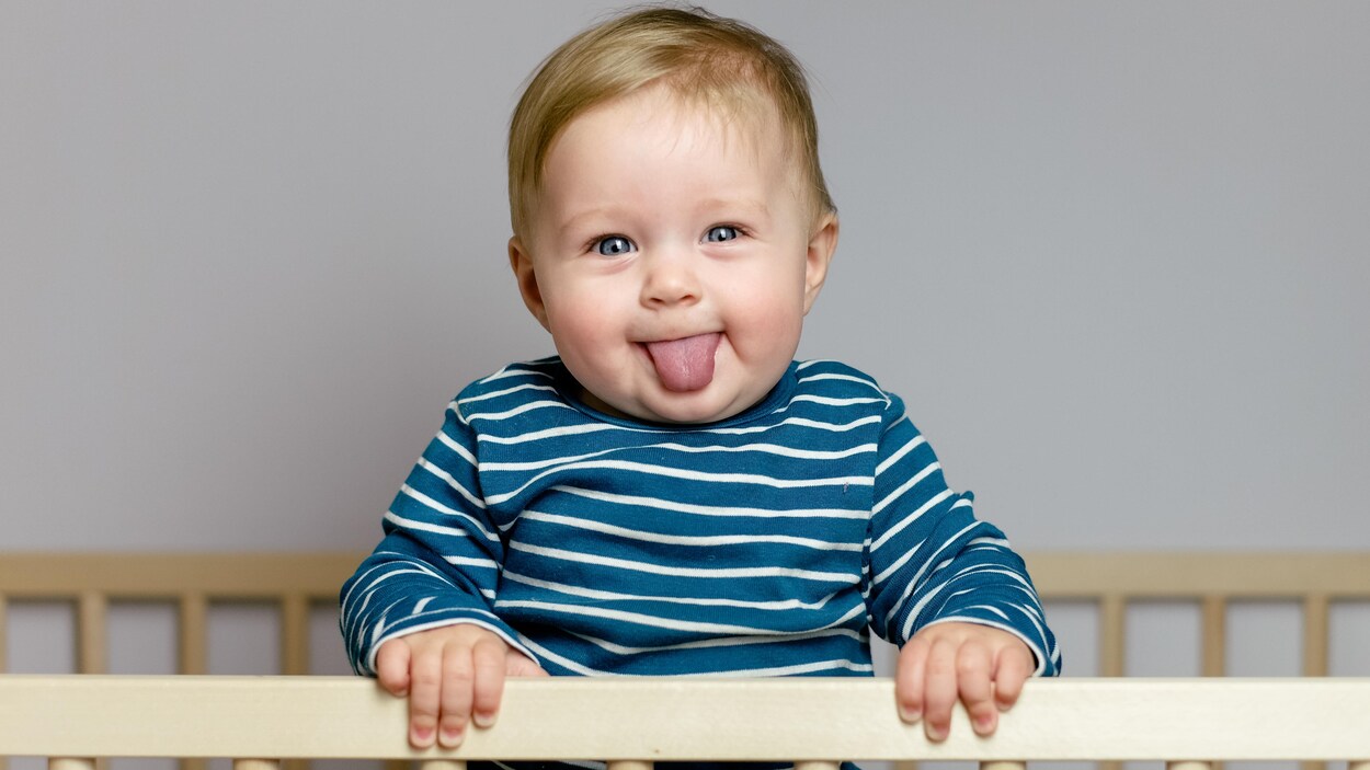 Un bébé debout dans son berceau qui grimace à la caméra Un bébé debout dans son berceau qui grimace à la caméra