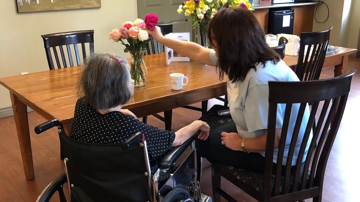 Une femme âgée en fauteuil roulant attablée à côté d'une femme qui prend une rose d'un vase posé sur la table.