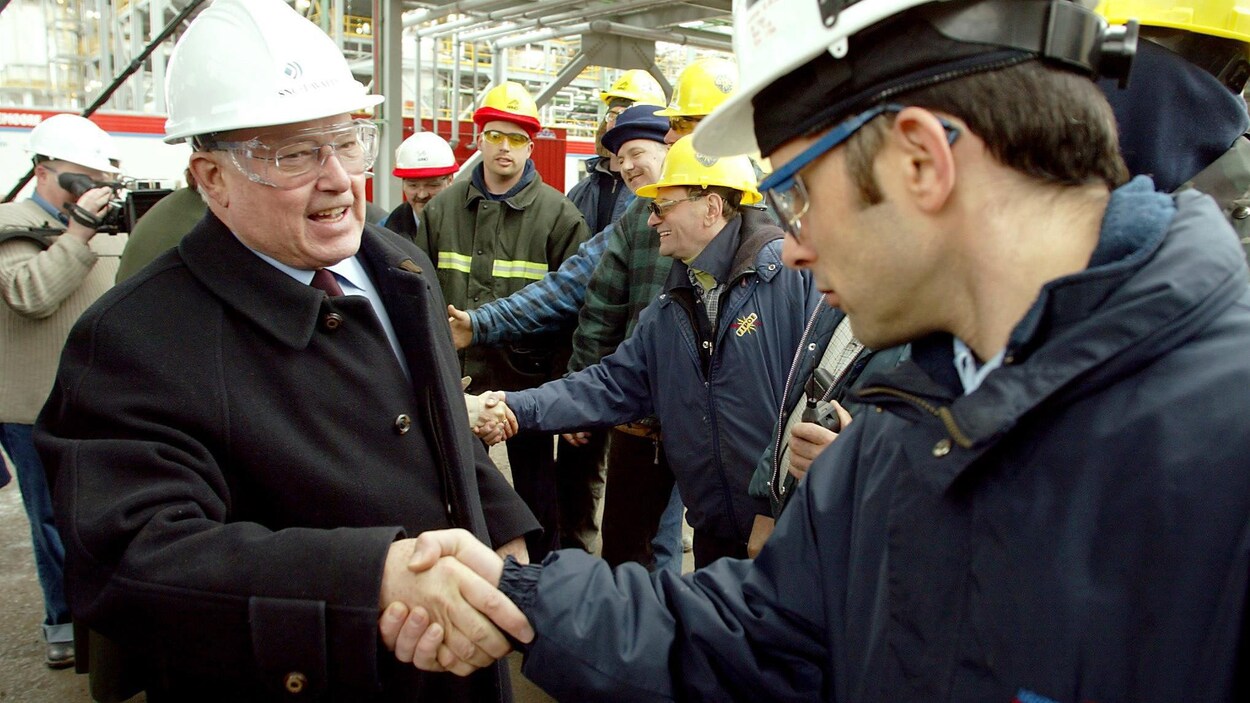 M. Landry, coiffé d'un casque de construction, serre la main d'un ouvrier en souriant.