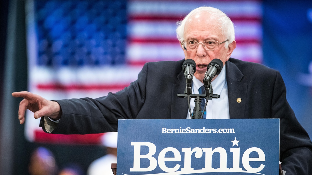 Un homme portant des lunettes parle derrière un lutrin en levant le bras droit. Une pancarte sur laquelle il est écrit «Bernie» est posée devant lui.