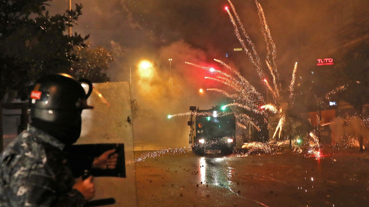 Un membre des forces de sécurité se protégeant avec un bouclier observe des feux d'artifice lancés quelques mètres devant lui.