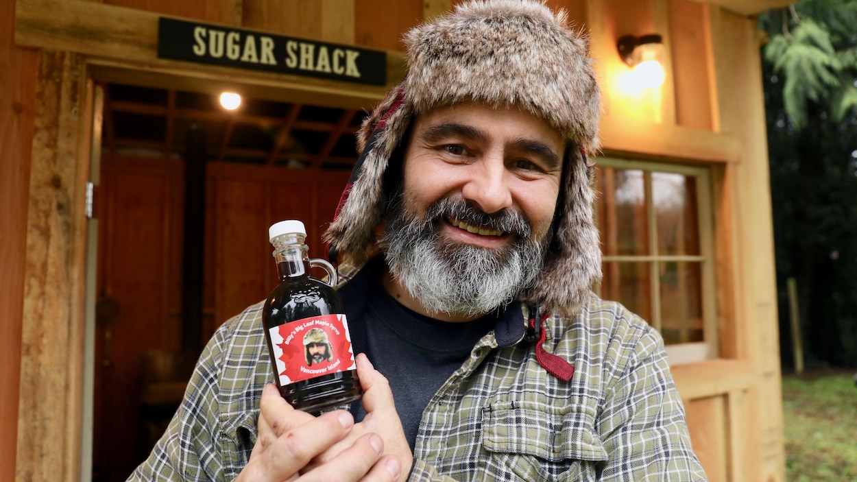 Un homme tient une bouteille de sirop d'érable devant sa cabane à sucre.