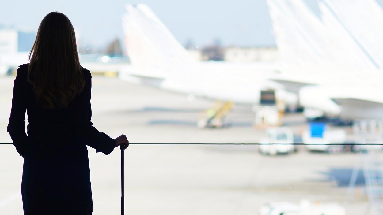 Une femme avec une valise roulante regarde des avions stationnés par la baie vitrée d'un terminal aéroportuaire.