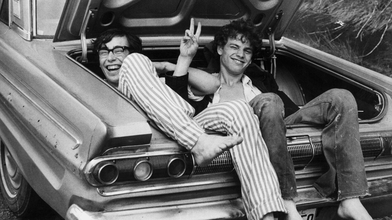 Deux jeunes hommes installés dans le coffre d'une voiture sourient à la caméra et l'un d'eux fait un signe de paix, après le festival de Woodstock, en août 1969.