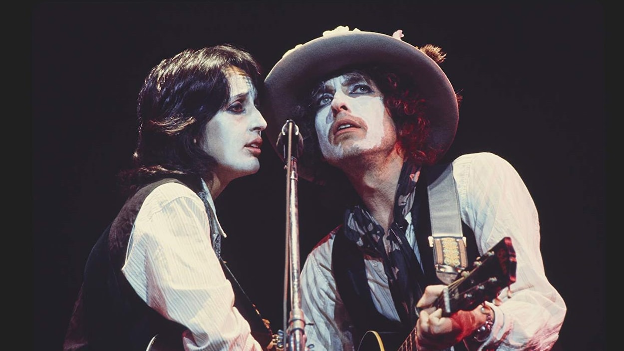 Une chanteuse et un chanteur sont sur scène devant un micro, guitare à la main. Ils ont le visage couvert de maquillage blanc.