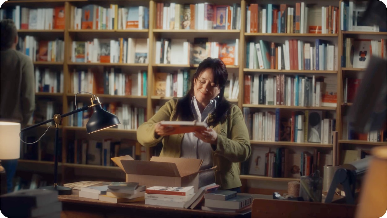 Une femme en gilet sort un livre d'un carton posé sur un comptoir encombré, devant une grande bibliothèque murale.