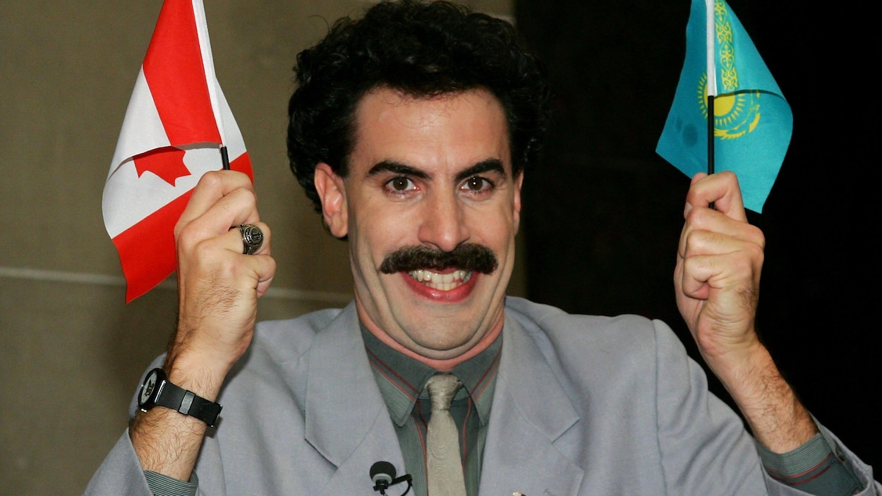 Un homme ayant une épaisse moustache tient un drapeau du Canada et un drapeau du Kazakhstan dans ses mains en souriant.