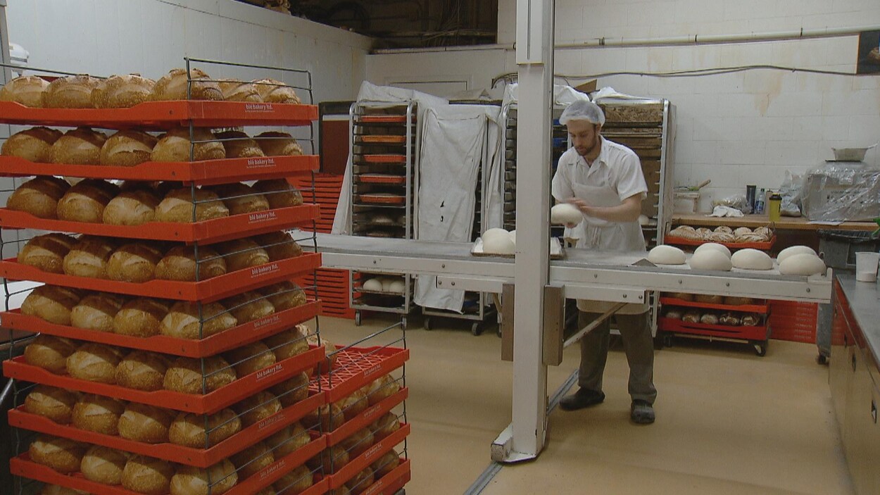 Un homme en uniforme de chef façonne de la farine sur un comptoir alors que des miches cuites reposent sur des barquettes.