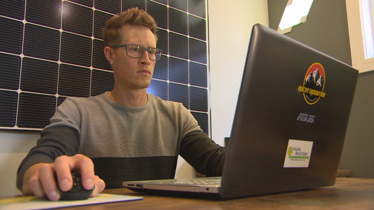 Dans un bureau, un homme l'air sérieux travaille avec un ordinateur portable avec le logo « Roots Rock Solar ». Derrière lui se trouve un panneau solaire.