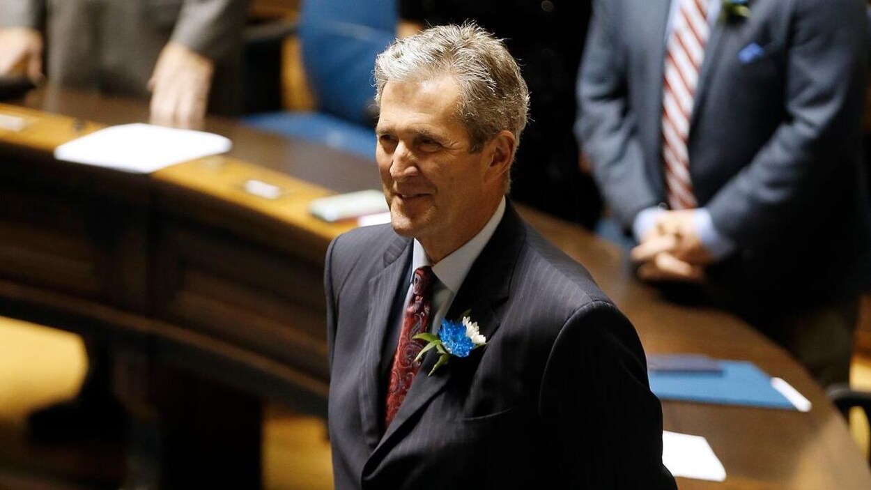 Brian Pallister est debout et souriant dans la Chambre de l'assemblée. On voit des députés également debout en arrière-plan.