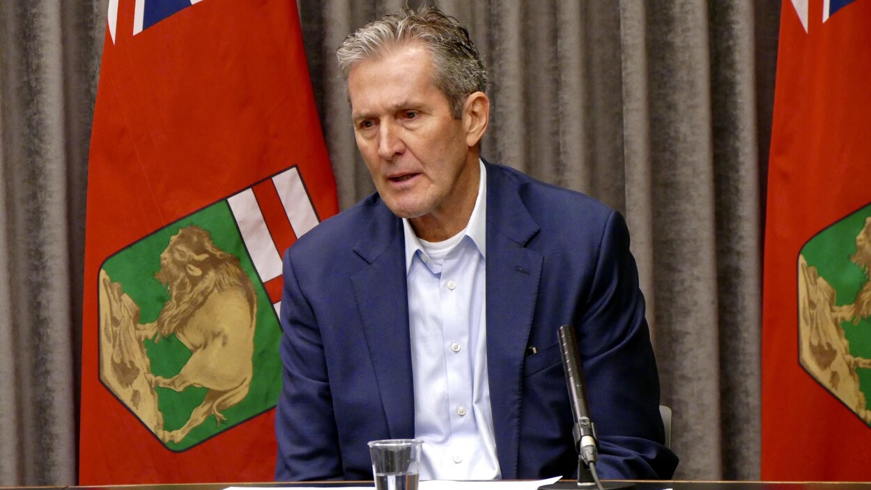 Le premier ministre du Manitoba, Brian Pallister, est assis à une table, avec derrière lui deux drapeaux de la province.