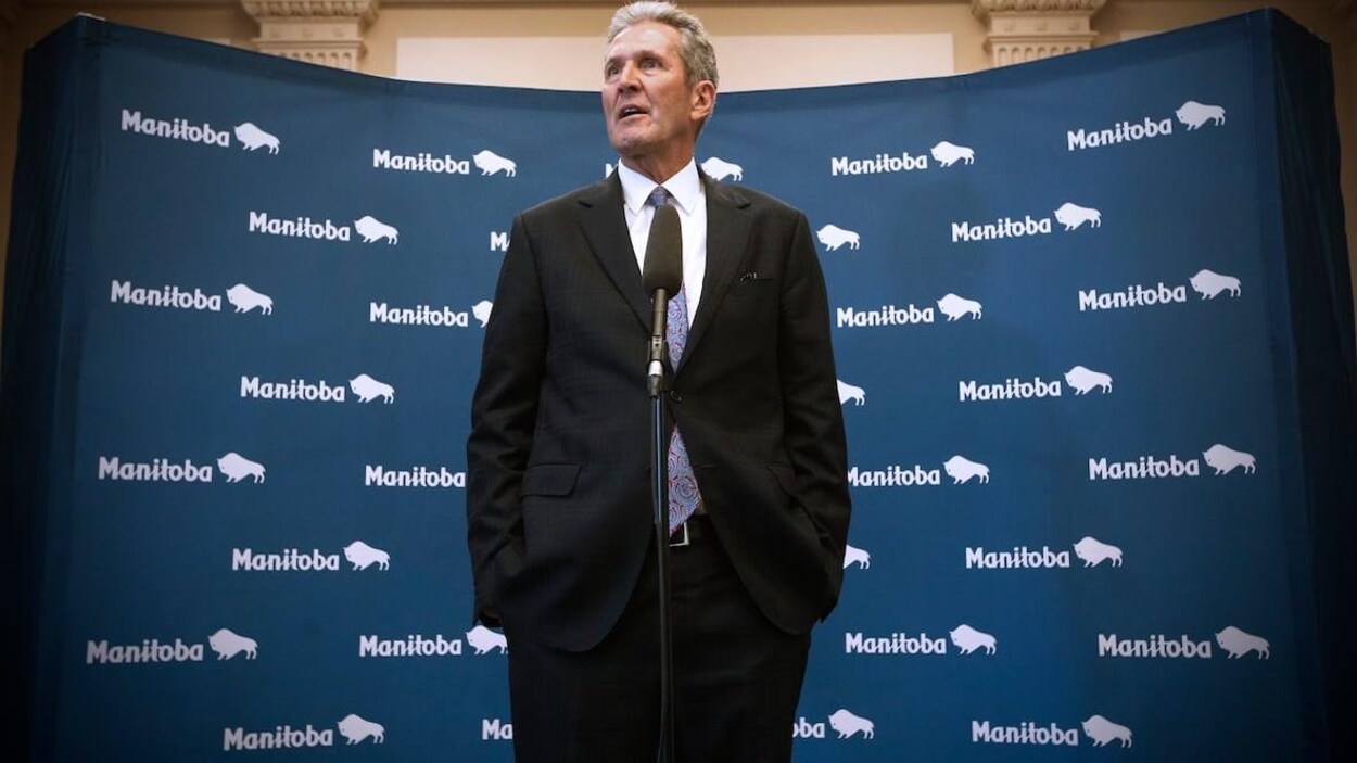 Brian Pallister parle, debout, derrière un micro et a les mains dans ses poches. On voit en arrière-plan plusieurs image du logo de la province (le mot Manitoba suivi d'une image stylisée de bison).