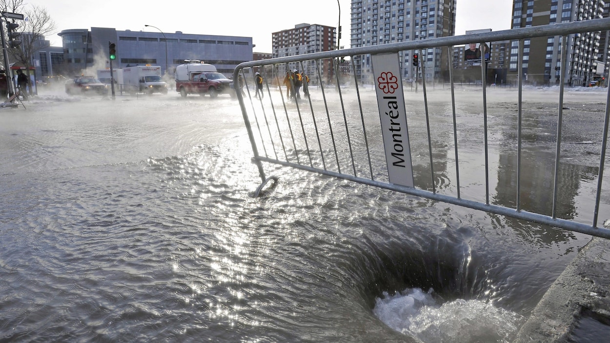 Vue panoramique d'une intersection inondée, une barrière métallique ornée du logo de la Ville de Montréal au premier plan.