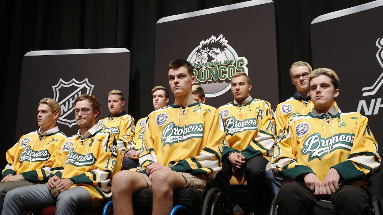 Les joueurs survivants se tiennent devant des affiches de la LNH et des Broncos de Humboldt. Ils portent tous des chandails de leur équipe de hockey.