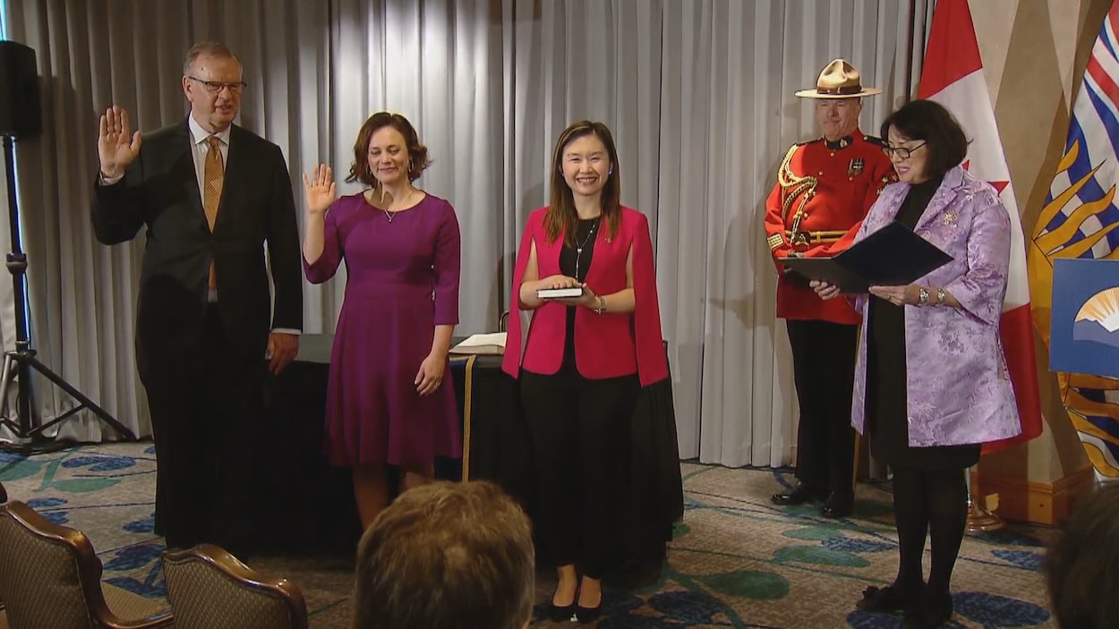 Bruce Ralston et Michelle Mungall récitent le serment en levant la main droite, tandis qu'Anne Kang sourit en gardant la main droite posée sur un livre. Janet Austin lit le serment sur un cahier. Derrière elle se trouvent un agent de la GRC en habit d'apparat et les drapeaux du Canada et de la Colombie-Britannique.