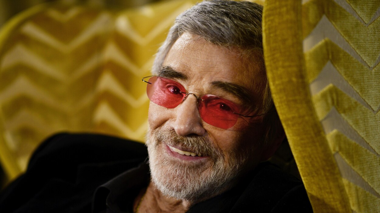 L'acteur Burt Reynolds est assis dans un fauteuil jaune et sourit à l'objectif.