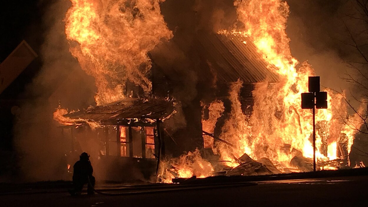 Un pompier combat un incendie. Une maison est complètement embrasée par d'imposantes flammes.