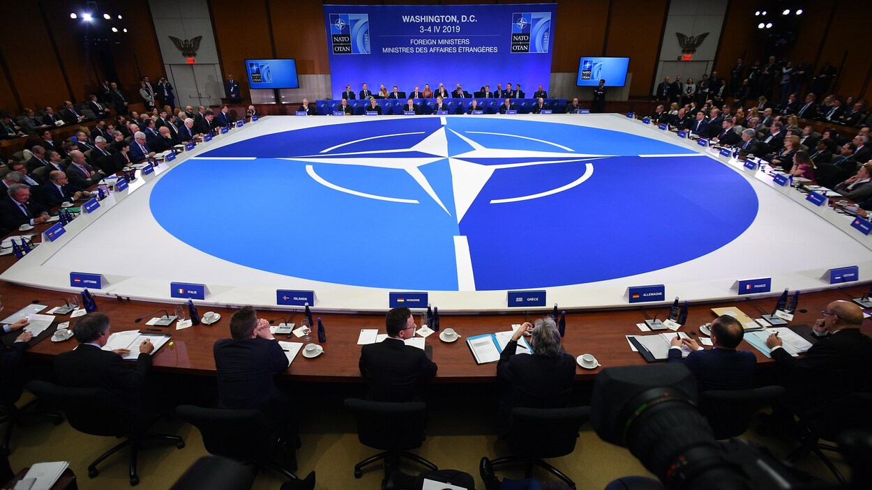 Des représentants des membres de l'OTAN assis à une énorme table sur laquelle le logo de l'organisation est mis à l'honneur.