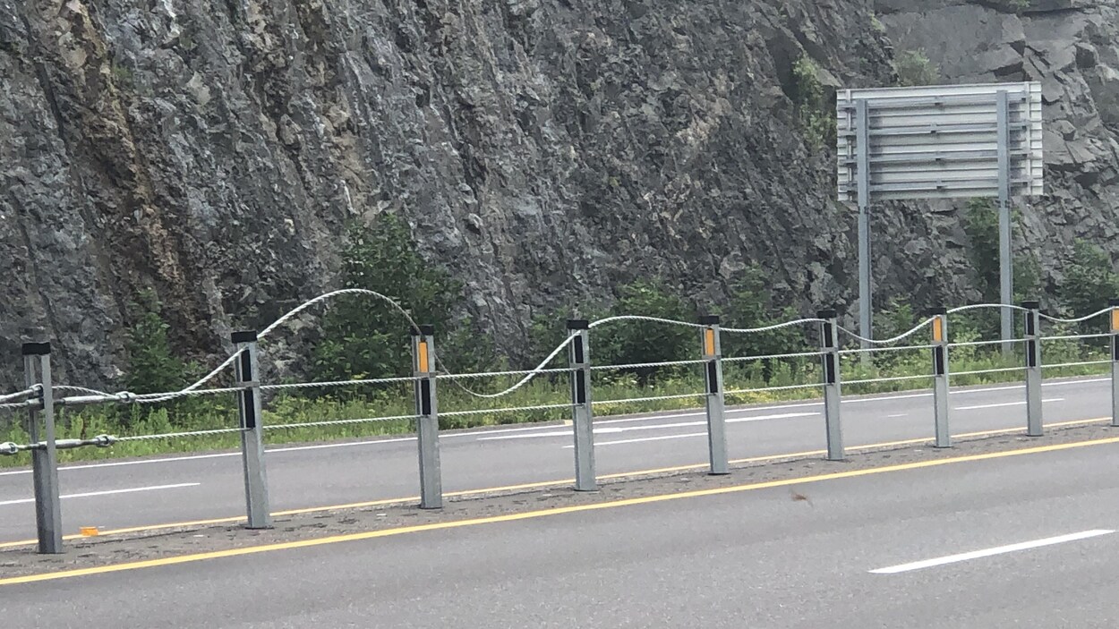 Une glissière composée de trois câbles de métal parallèles sur une route. Le câble du haut est sinueux, tandis que les deux autres sont tendus et droits.