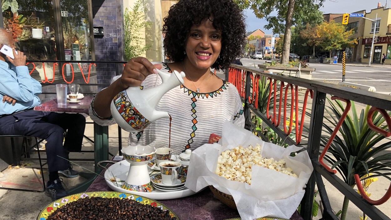 Une femme assise à la terrasse devant un homme qui parle au téléphone. Jebena à la main, elle verse le café dans des petites tasses rangées sur un plateau à coté de grains de café et de maïs soufflé.