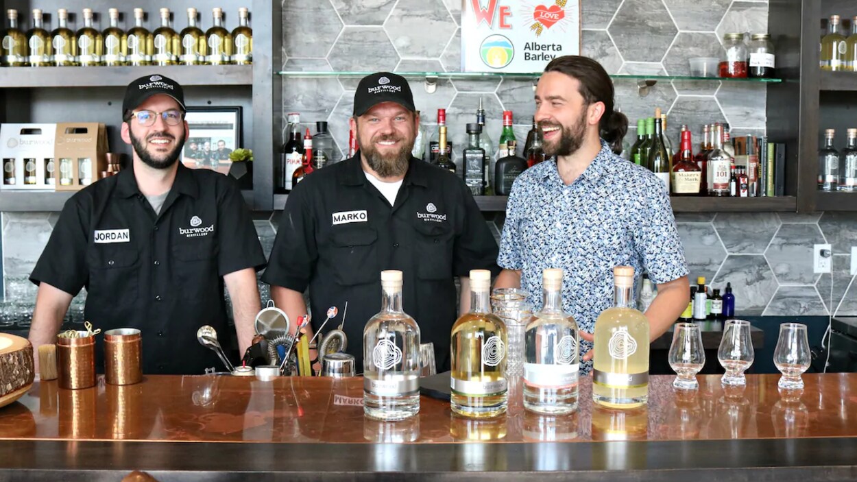 Jordan Ramey, à gauche, Marko Ramey et Ivan Cilic posent derrière un comptoir sur lequel trônent des bouteilles d'alcool de leur distillerie.