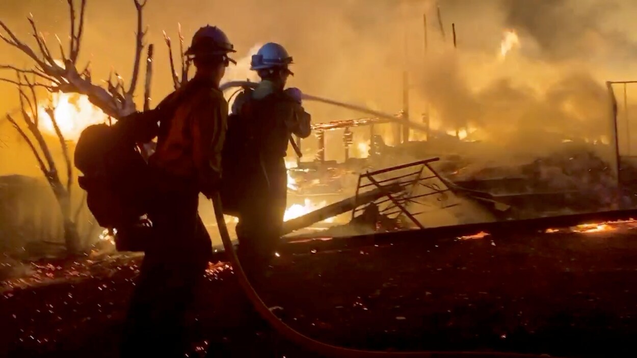 Deux pompiers tiennent une lance d'incendie à côté d'une maison détruite.