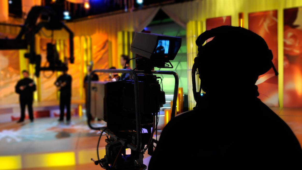 Une personne portant un casque d'écoute se tient derrière une caméra, alors qu'elle filme le plateau d'une émission de télévision.