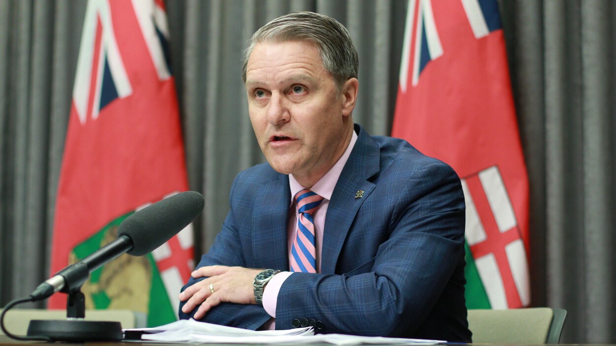 Un homme à une table avec un micro, et des drapeaux de la province du Manitoba derrière lui.