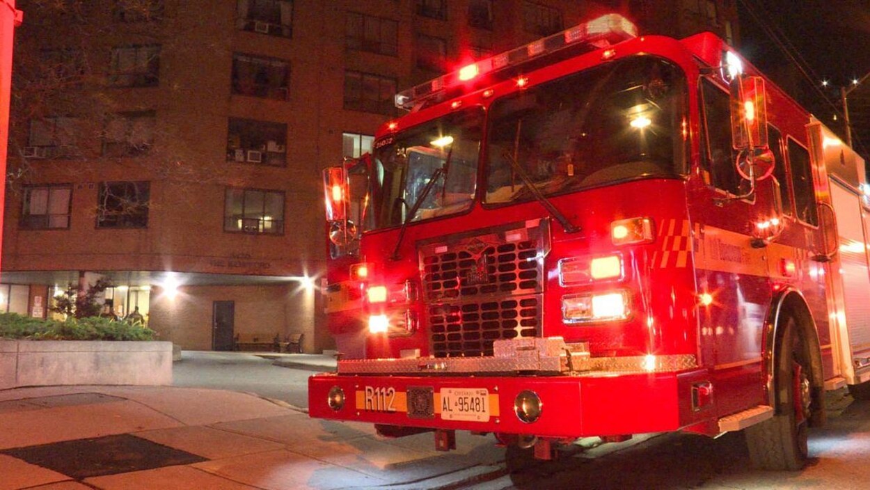 Photo d'un camion de pompier rouge aux lumières clignotantes devant un immeuble en briques brunes, le soir.