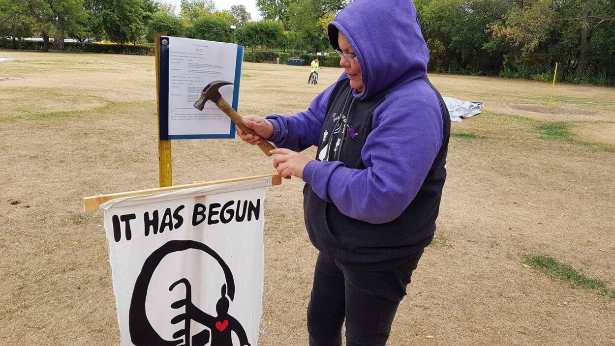 Une femme autochtone frappe avec un marteau sur une affiche où il est écrit « cela a commencé », en anglais. Une femme autochtone frappe avec un marteau sur une affiche où il est écrit « cela a commencé », en anglais.