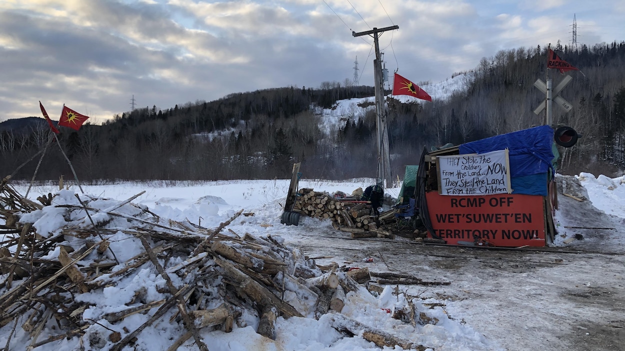 Un campement construit de planches et de baches, entouré de bois de chauffage et de drapeaux mohawks