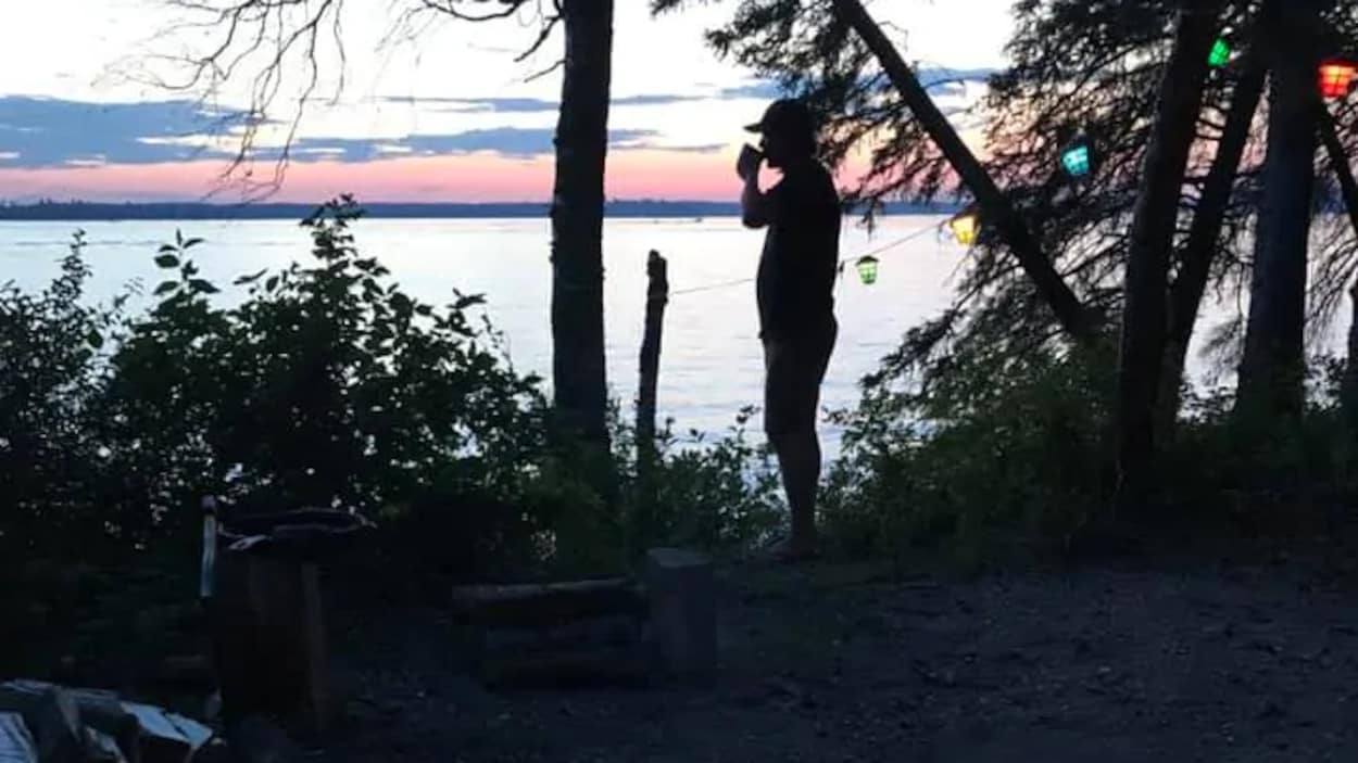 Une personne dans l'ombre regarde le coucher du soleil dans un terrain de camping sur le bord de l'eau. Un feu de camp est allumé.
