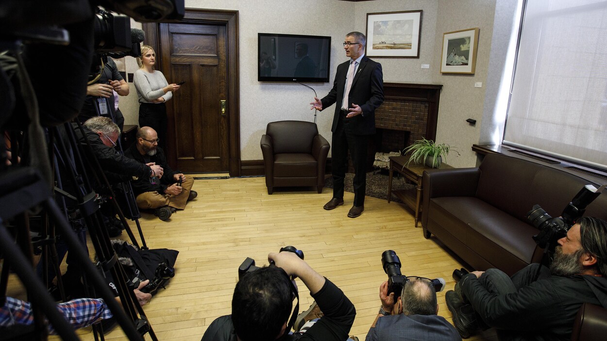 Travis Toews, debout dans son bureau, chaussé de bottes. Des photographes et des caméramans se pressent autour de lui.