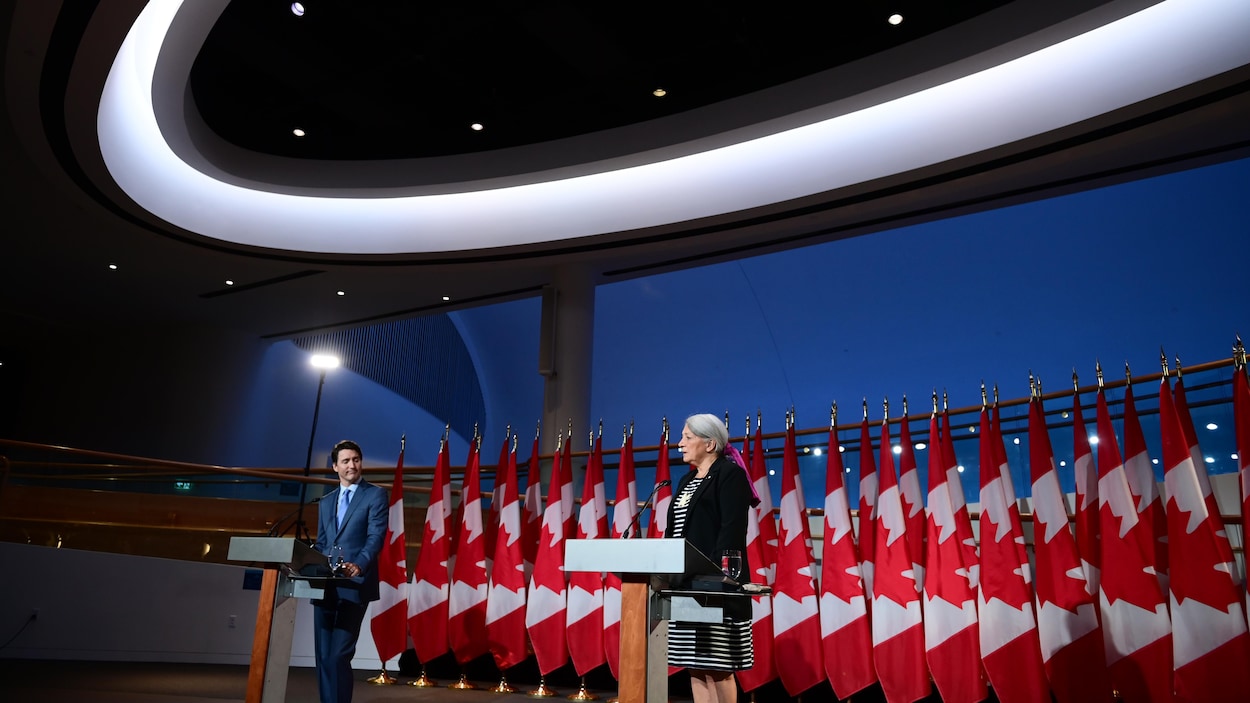 Mary Simon et Justin Trudeau, debout, devant une rangée de drapeaux du Canada, dans une salle éclairée par des projecteurs.