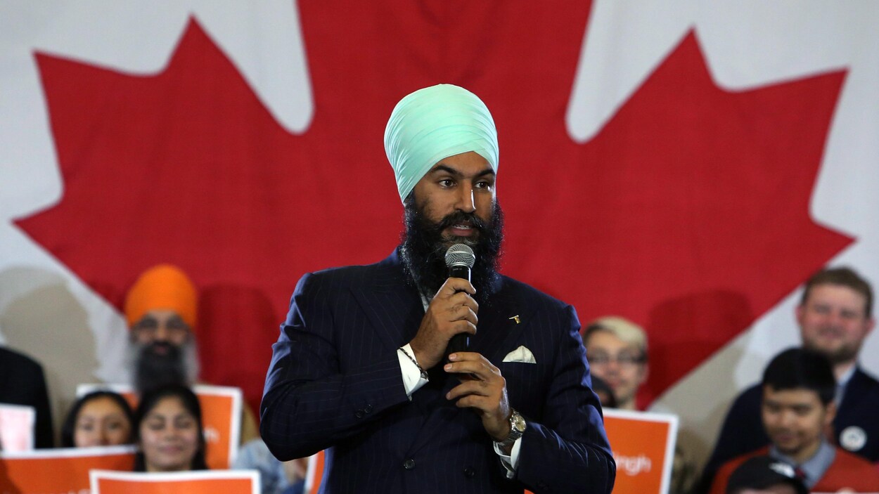 Un homme tient un micro devant un drapeau du Canada et des gens tenant des affiches orange.