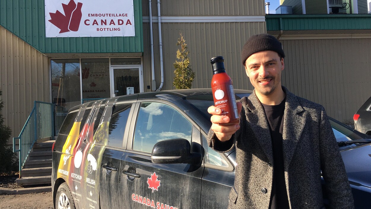 Un homme tient une bouteille de ketchup devant un véhicule stationné devant un commerce d'embouteillage.