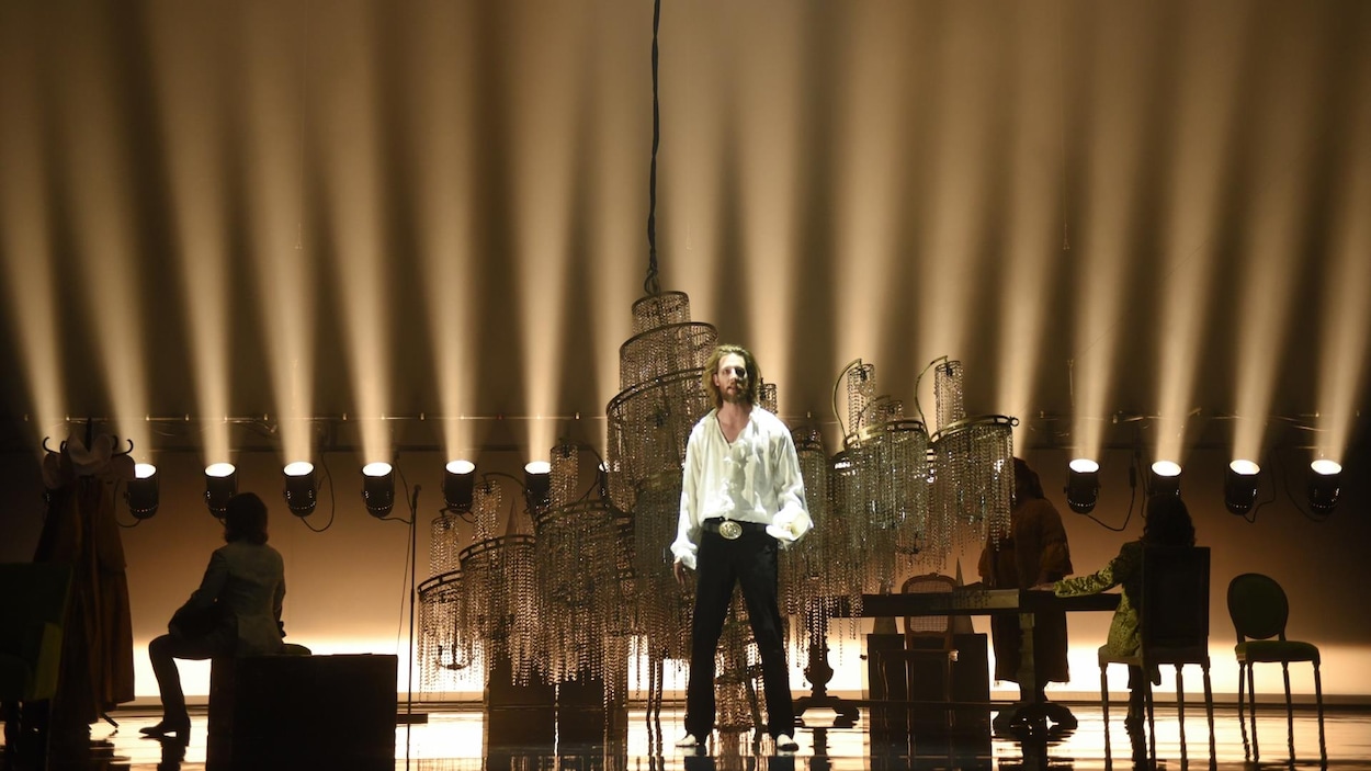 Emmanuel Schwartz se tient debout derrière un lustre géant. On aperçoit également d'autres acteurs de dos, dans l'ombre, assis sur une chaise ou sur un siège. Emmanuel Schwartz se tient debout derrière un lustre géant. On aperçoit également d'autres acteurs de dos, dans l'ombre, assis sur une chaise ou sur un siège.