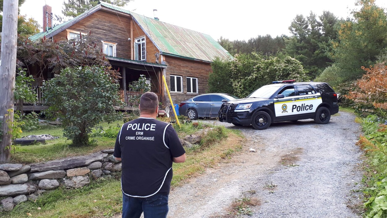 Un policier de dos en premier plan avec devant lui, deux voitures dont une de la Sûreté du Québec et une maison.