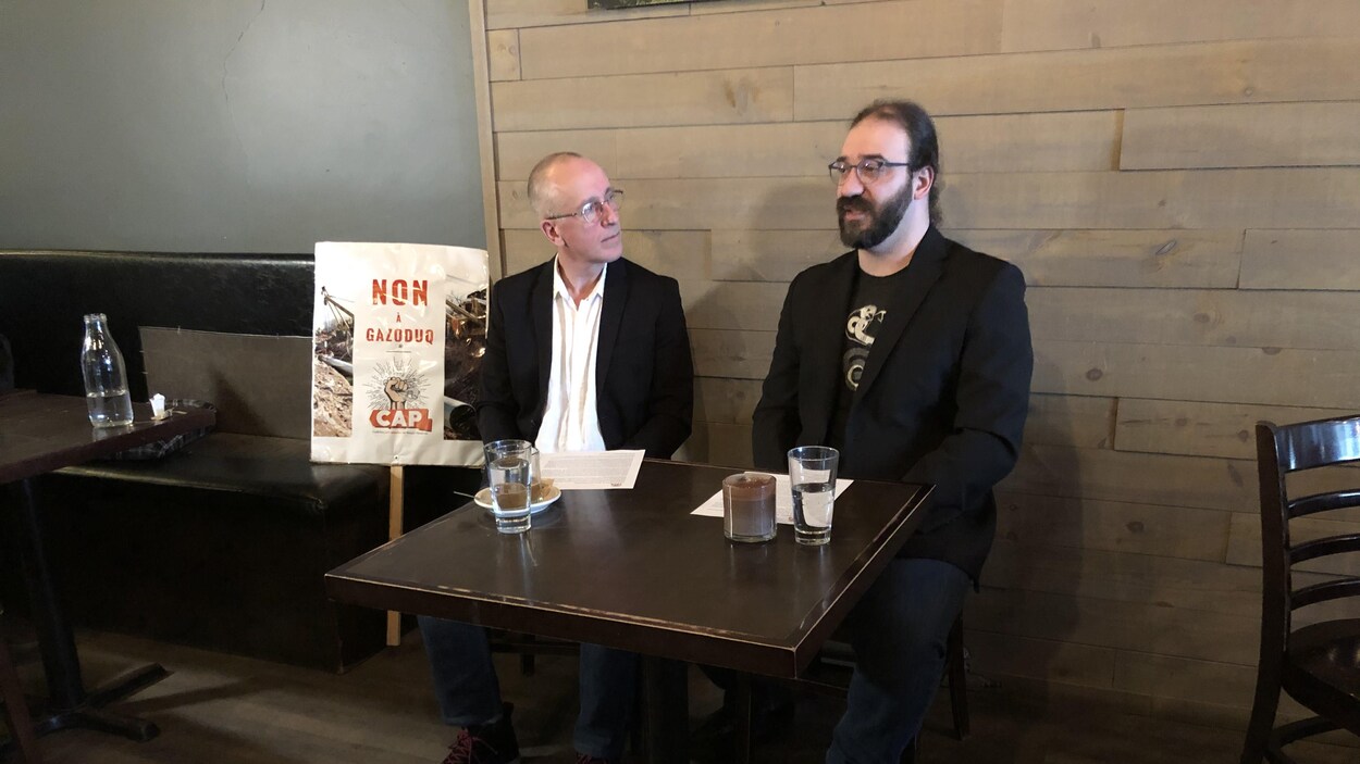 Deux hommes parlent dans un restaurant. Une pancarte indiquant « Non à Gazoduq » est placée près d'eux.