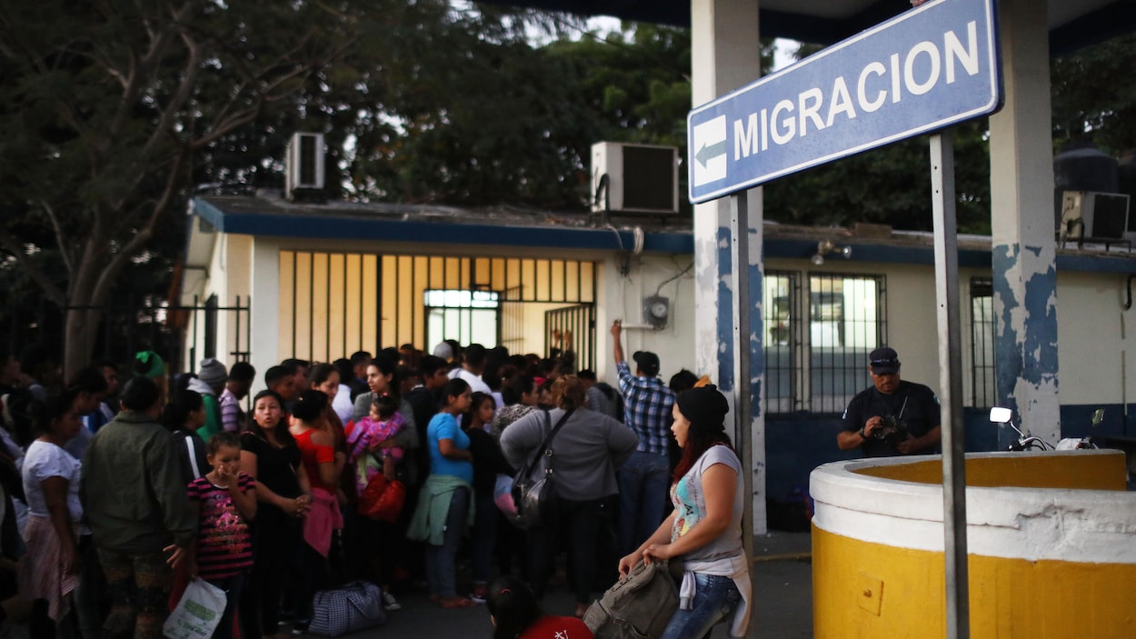 Des personnes font la queue derrière une barrière. Un panneau indique « Migration ».