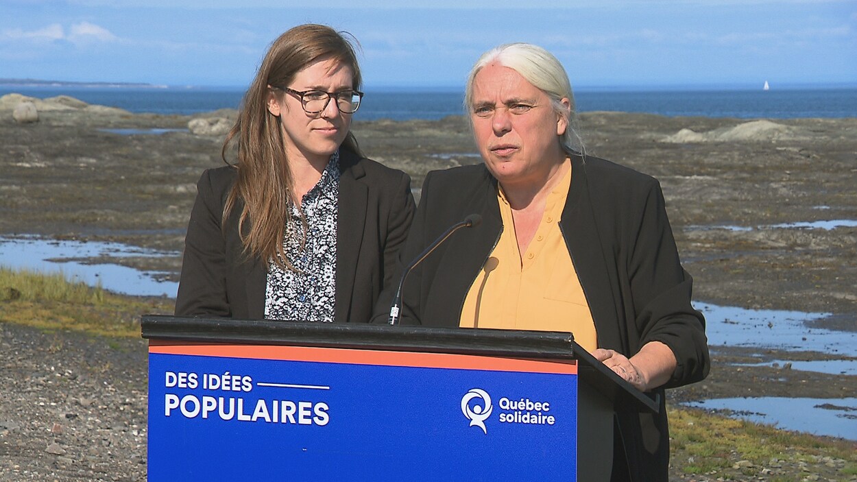 Installée sur la grève, Manon Massé parle au micro, derrière un lutrin aux couleurs de Québec solidaire. Carol-Ann Kack la regarde. En arrière-plan, le fleuve et un voilier au loin.