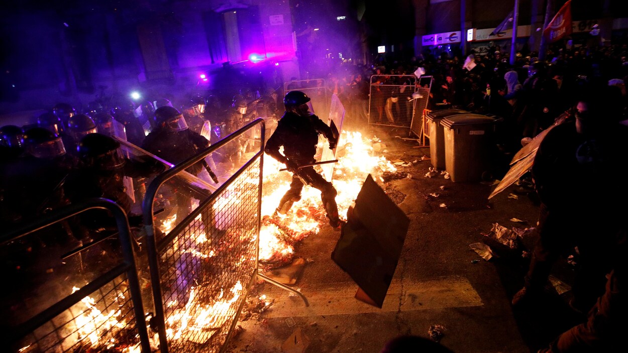 Des policiers masqués et munis de matraques traversent une ligne de cartons enflammés pour affronter les manifestants.