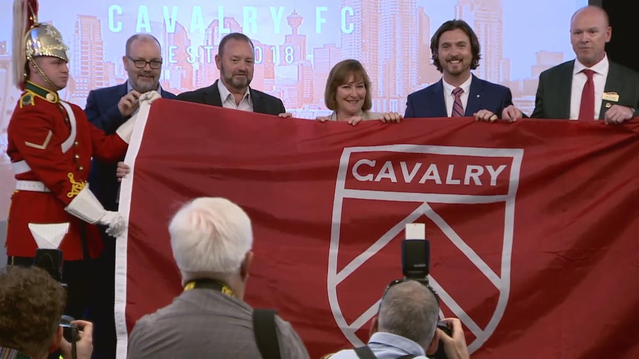 Des personnes tiennent un grand drapeau qui porte le blason de la nouvelle équipe de soccer de Calgary, devant les photographes. Des personnes tiennent un grand drapeau qui porte le blason de la nouvelle équipe de soccer de Calgary, devant les photographes.