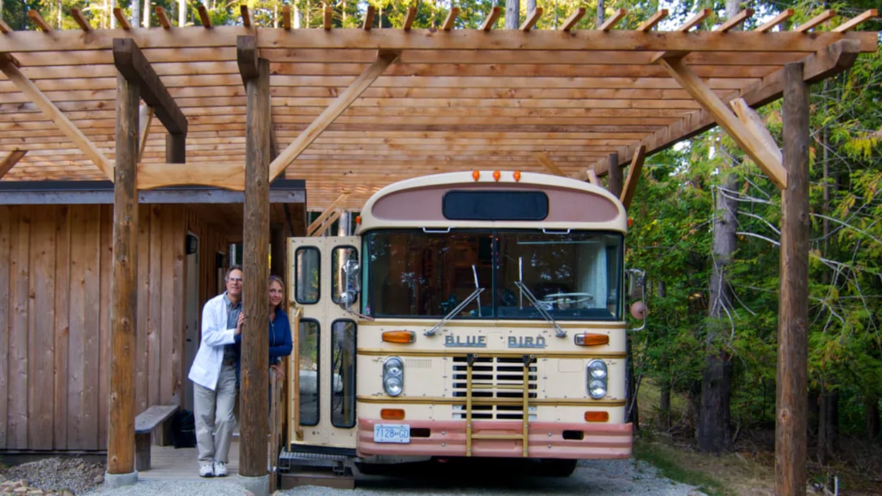 Un homme en sarrau et une femme se tiennent à côté d'un bus, sous une pergola de bois.