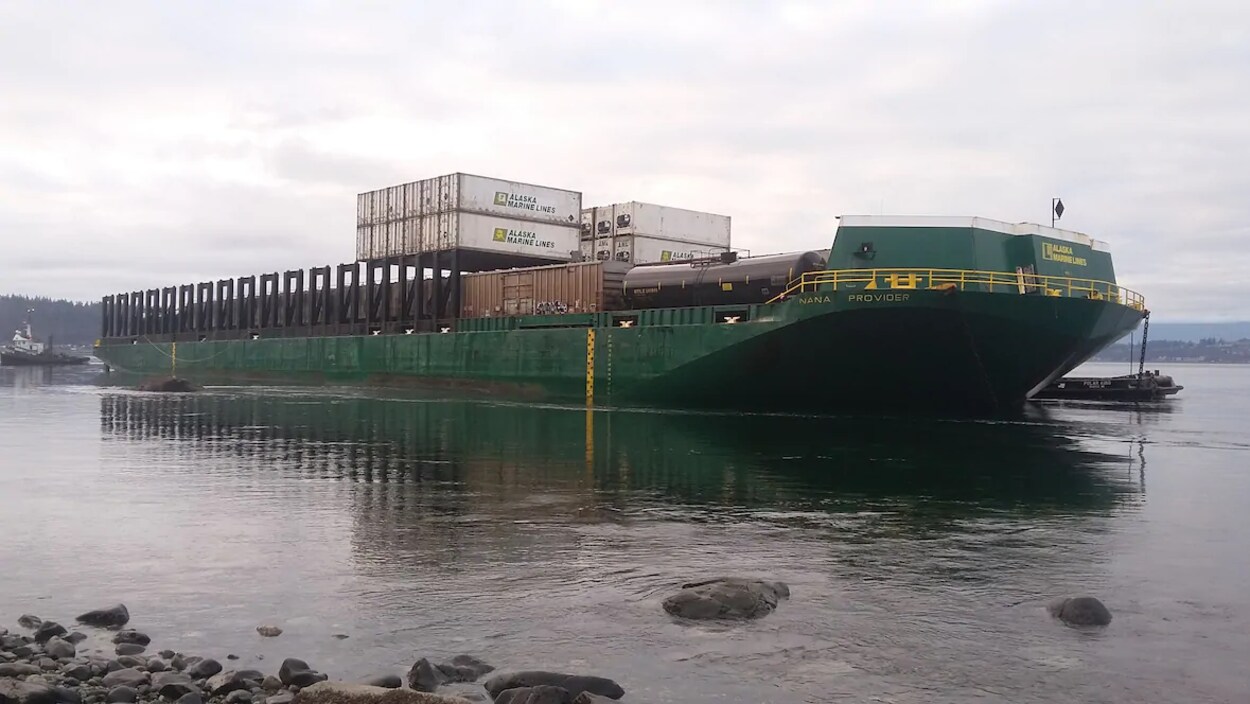 Une barge au bord d'une plage, entourée de 2 remorqueurs.