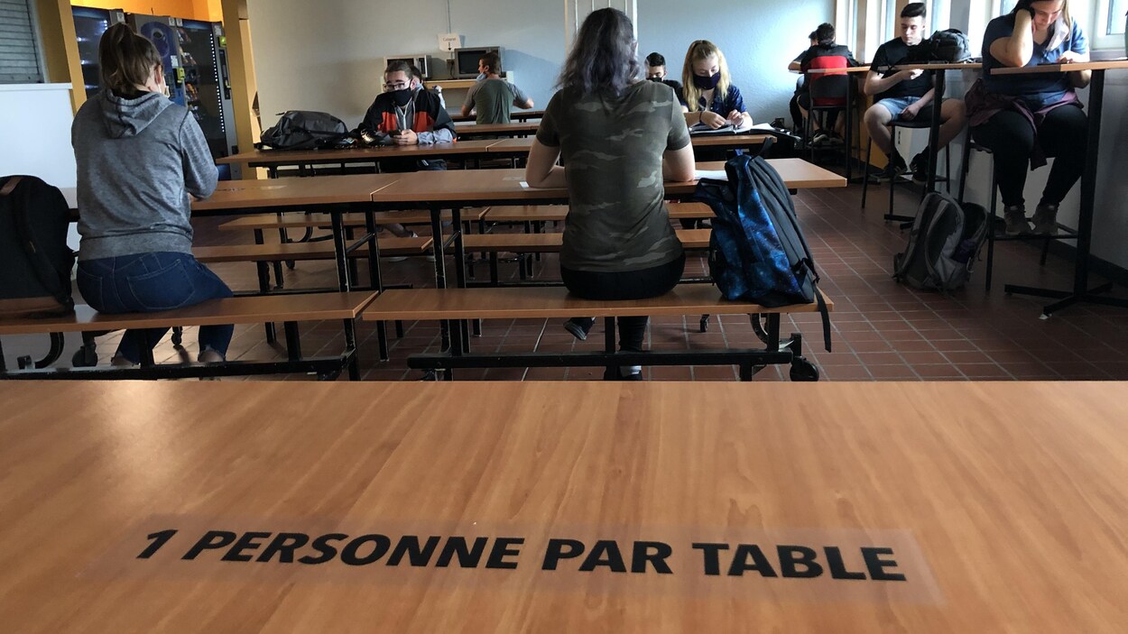 Une table où il est inscrit: "1 personne par table". On voit des jeunes assis en arrière plan.